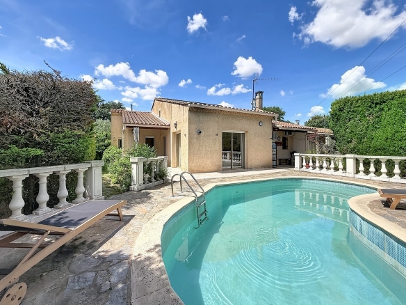 Piscine arrondie avec eau turquoise entourée d'une terrasse en pierre et balustrades blanches. Façade de maison provençale enduit ocre avec toiture en tuiles et transats en bois.