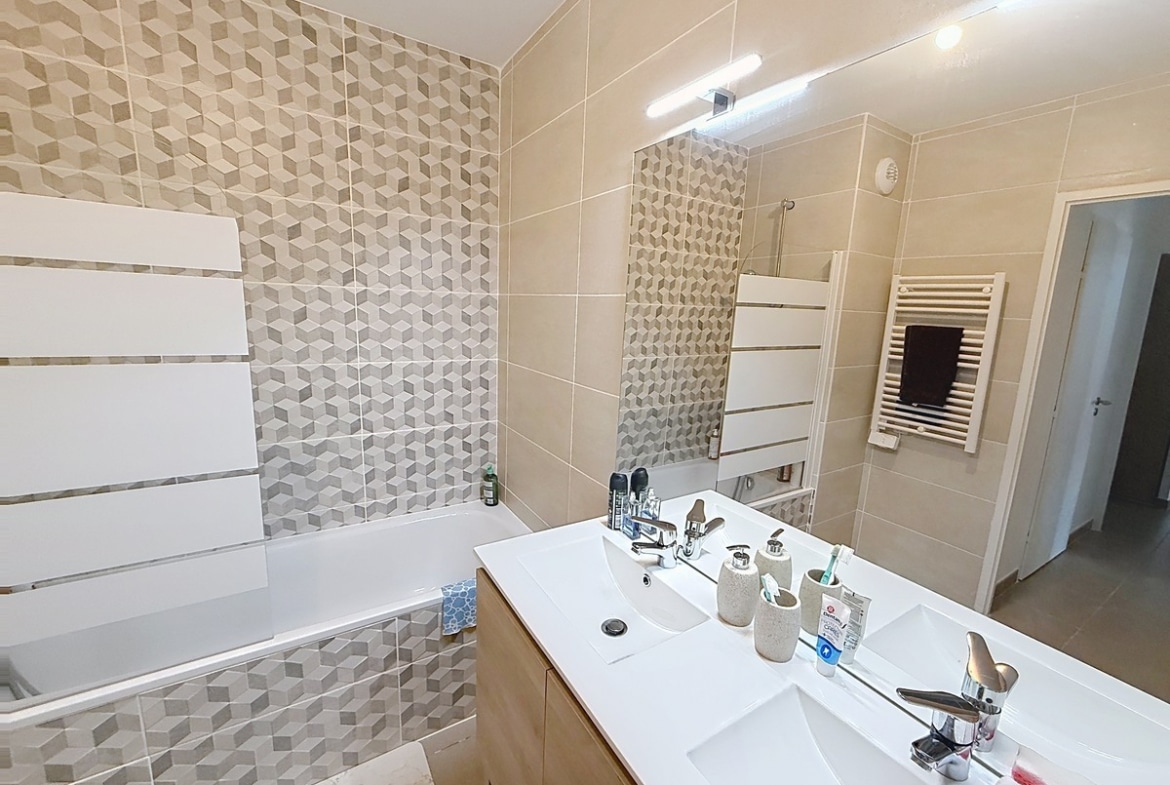 Salle de bains carrelée en tons beige avec mur d'accent à motifs géométriques 3D gris et beige. Double vasque blanche sur meuble en bois clair, baignoire encastrée et sèche-serviettes blanc mural.