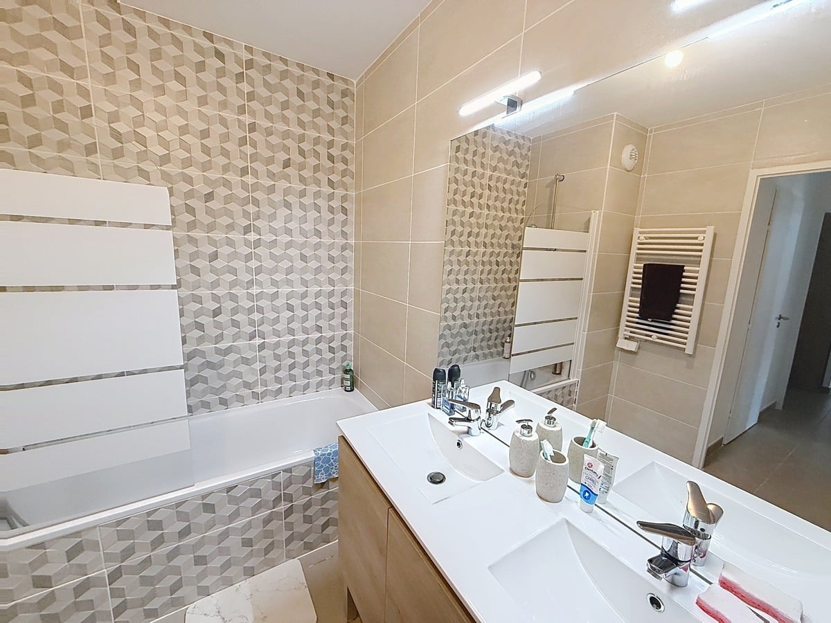 Salle de bains carrelée en tons beige avec mur d'accent à motifs géométriques 3D gris et beige. Double vasque blanche sur meuble en bois clair, baignoire encastrée et sèche-serviettes blanc mural.
