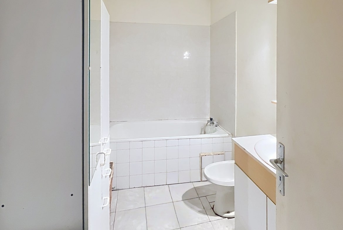 Salle de bains avec carrelage blanc au sol et faïence blanche murale autour de la baignoire encastrée. Un meuble vasque en bois clair avec lavabo rond blanc est installé à droite, et un grand miroir occupe le mur au-dessus.