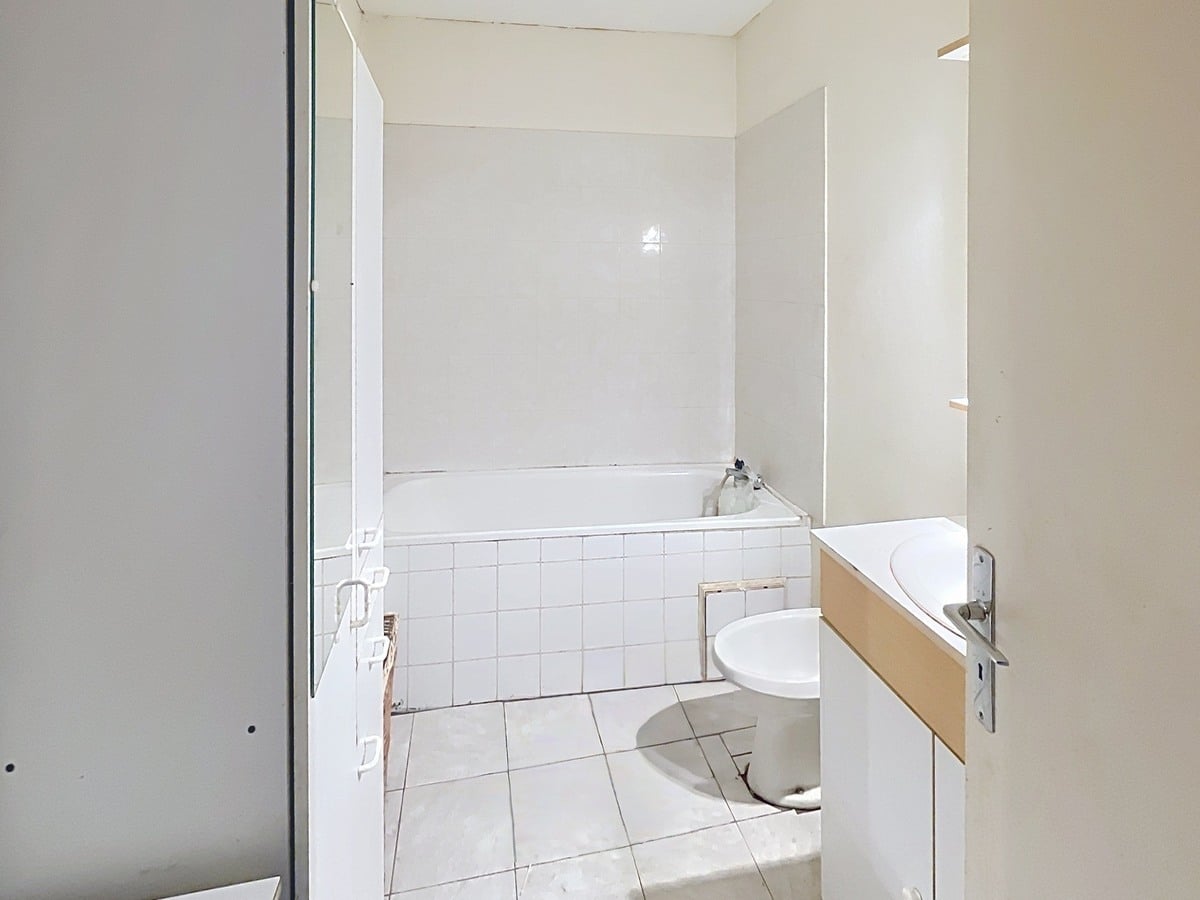 Salle de bains avec carrelage blanc au sol et faïence blanche murale autour de la baignoire encastrée. Un meuble vasque en bois clair avec lavabo rond blanc est installé à droite, et un grand miroir occupe le mur au-dessus.