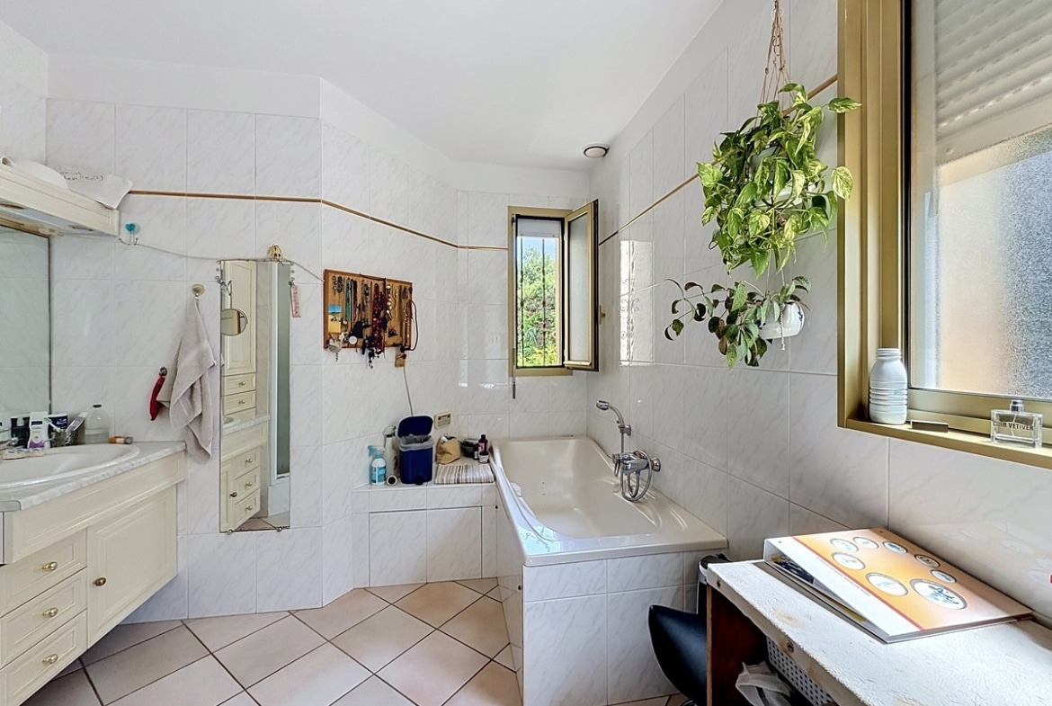 Salle de bains carrelée blanc avec baignoire encastrée et meuble vasque crème. Deux fenêtres avec cadre doré, plante suspendue verte et sol en carrelage beige diagonal.