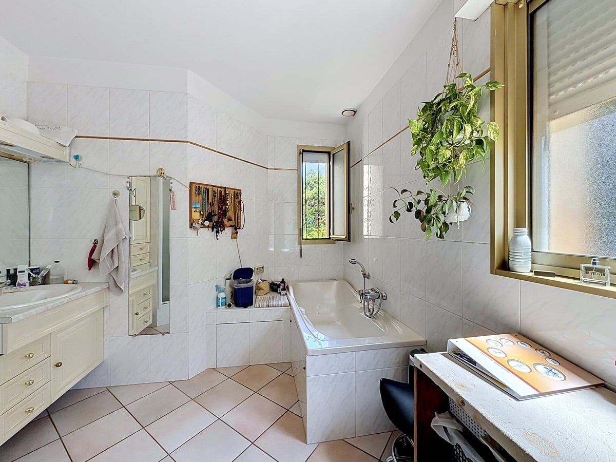 Salle de bains carrelée blanc avec baignoire encastrée et meuble vasque crème. Deux fenêtres avec cadre doré, plante suspendue verte et sol en carrelage beige diagonal.