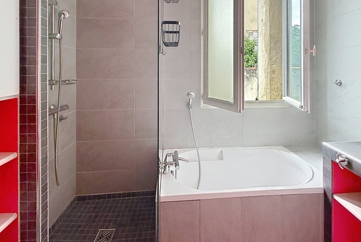 Salle de bains carrelée gris avec douche à l'italienne au sol en mosaïque noire et baignoire encastrée. Meubles de rangement rouges de chaque côté, fenêtre à battants ouverte sur l'extérieur.