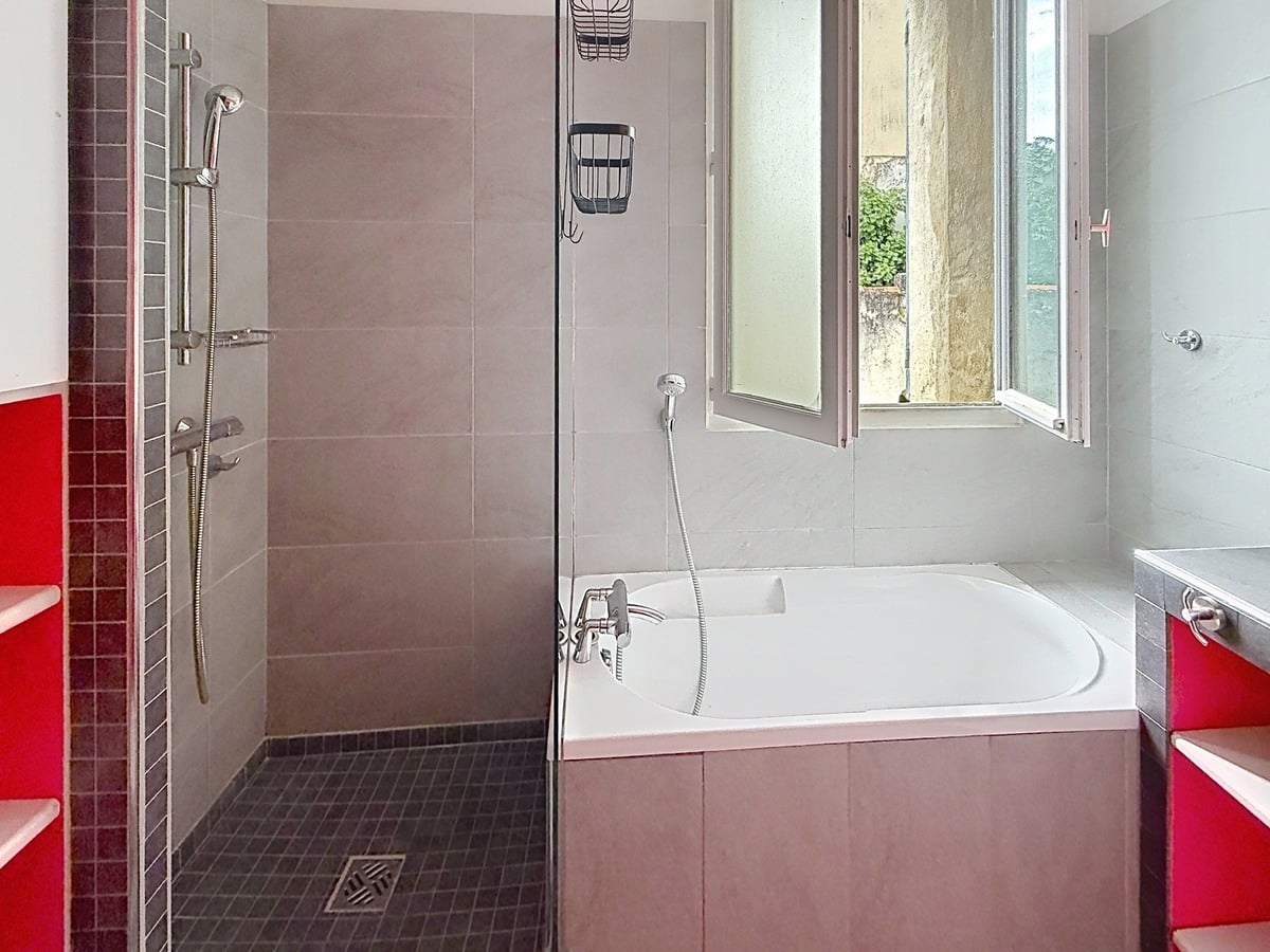 Salle de bains carrelée gris avec douche à l'italienne au sol en mosaïque noire et baignoire encastrée. Meubles de rangement rouges de chaque côté, fenêtre à battants ouverte sur l'extérieur.