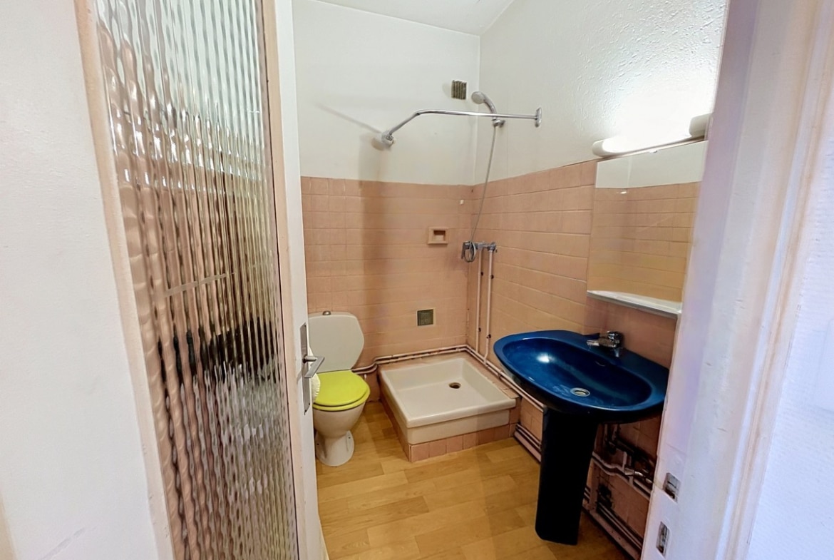 Salle de bains avec carrelage mural rose pêche, receveur de douche blanc surélevé et douchette chromée. Lavabo bleu sur pied noir, WC avec abattant jaune, sol en vinyle imitation bois, porte vitrée texturée.