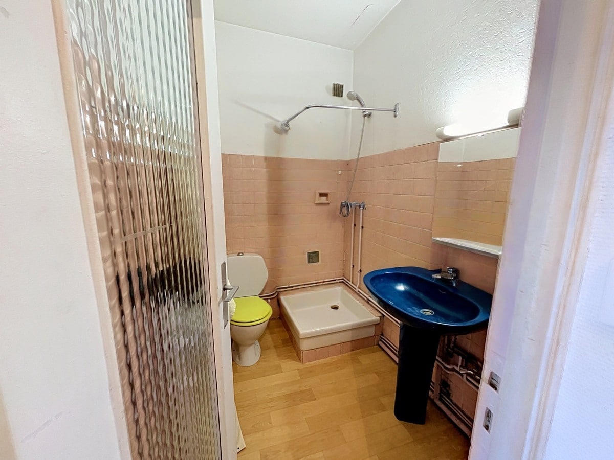 Salle de bains avec carrelage mural rose pêche, receveur de douche blanc surélevé et douchette chromée. Lavabo bleu sur pied noir, WC avec abattant jaune, sol en vinyle imitation bois, porte vitrée texturée.