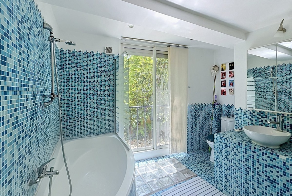 Salle de bains entièrement carrelée en mosaïque bleue et blanche avec baignoire d'angle, douche et toilettes intégrés. Vasque posée sur un meuble carrelé, fenêtre coulissante donnant sur végétation extérieure et pavés de verre au sol.