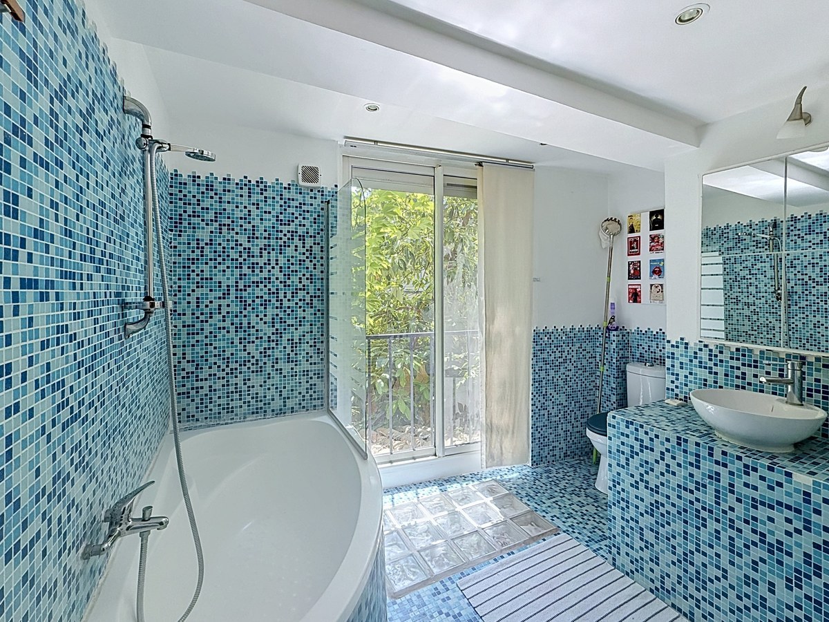 Salle de bains entièrement carrelée en mosaïque bleue et blanche avec baignoire d'angle, douche et toilettes intégrés. Vasque posée sur un meuble carrelé, fenêtre coulissante donnant sur végétation extérieure et pavés de verre au sol.