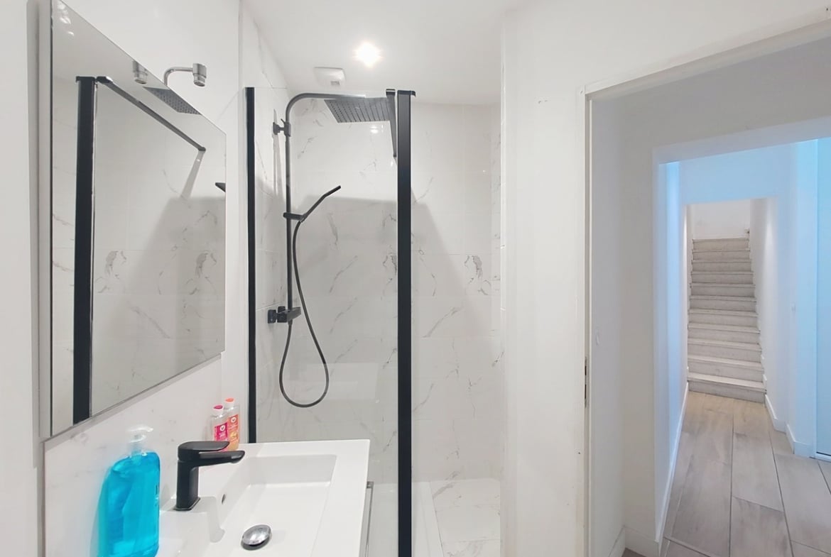 Salle d'eau moderne avec carrelage mural effet marbre blanc veiné gris. La douche est équipée d'une paroi vitrée à cadre noir et d'une colonne de douche noire. Un meuble vasque blanc avec robinetterie noire fait face à un miroir rectangulaire.