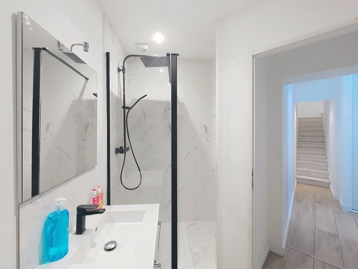 Salle d'eau moderne avec carrelage mural effet marbre blanc veiné gris. La douche est équipée d'une paroi vitrée à cadre noir et d'une colonne de douche noire. Un meuble vasque blanc avec robinetterie noire fait face à un miroir rectangulaire.