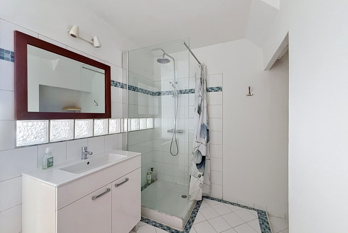 Salle d'eau avec meuble vasque blanc, miroir à cadre bordeaux et douche à l'italienne avec paroi vitrée. Carrelage blanc au mur avec frise en mosaïque bleue, sol carrelé blanc avec motifs géométriques bleus.