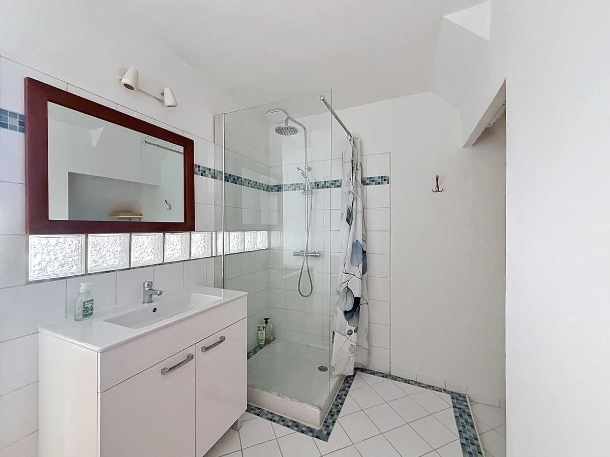 Salle d'eau avec meuble vasque blanc, miroir à cadre bordeaux et douche à l'italienne avec paroi vitrée. Carrelage blanc au mur avec frise en mosaïque bleue, sol carrelé blanc avec motifs géométriques bleus.