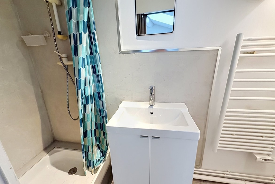 Petite salle d'eau blanche avec douche équipée d'un rideau à motifs géométriques bleus. Meuble vasque blanc avec miroir, sèche-serviettes mural blanc, fenêtre obscure en partie haute.