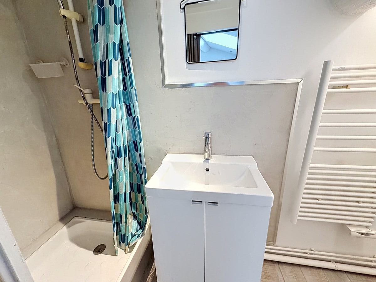 Petite salle d'eau blanche avec douche équipée d'un rideau à motifs géométriques bleus. Meuble vasque blanc avec miroir, sèche-serviettes mural blanc, fenêtre obscure en partie haute.