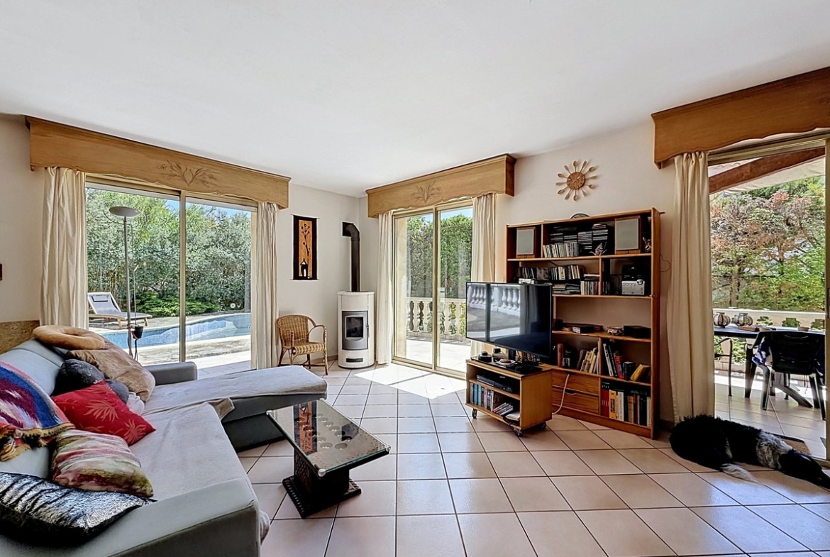 Salon carrelé beige avec canapé gris orné de coussins colorés et poêle à bois blanc. Bibliothèque en bois, télévision et doubles baies vitrées donnant sur la piscine et la terrasse.