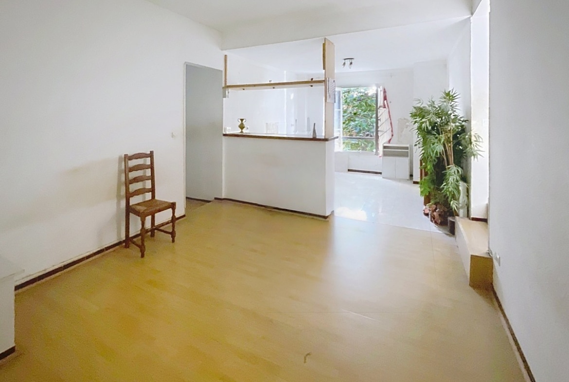 Séjour vide avec parquet clair et cuisine ouverte séparée par un bar en bois avec étagères. Un escalier rouge est visible à droite près d'une grande plante verte et d'une fenêtre donnant sur la végétation extérieure.