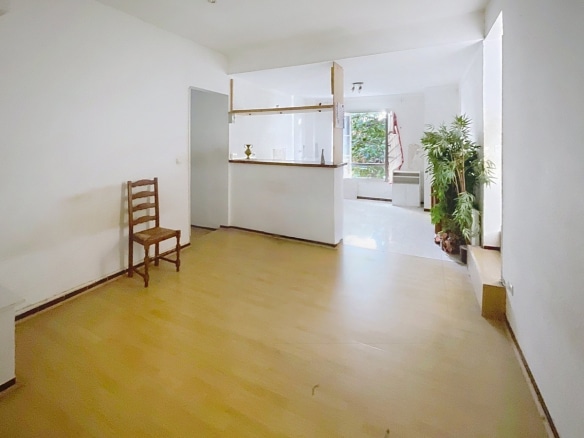 Séjour vide avec parquet clair et cuisine ouverte séparée par un bar en bois avec étagères. Un escalier rouge est visible à droite près d'une grande plante verte et d'une fenêtre donnant sur la végétation extérieure.