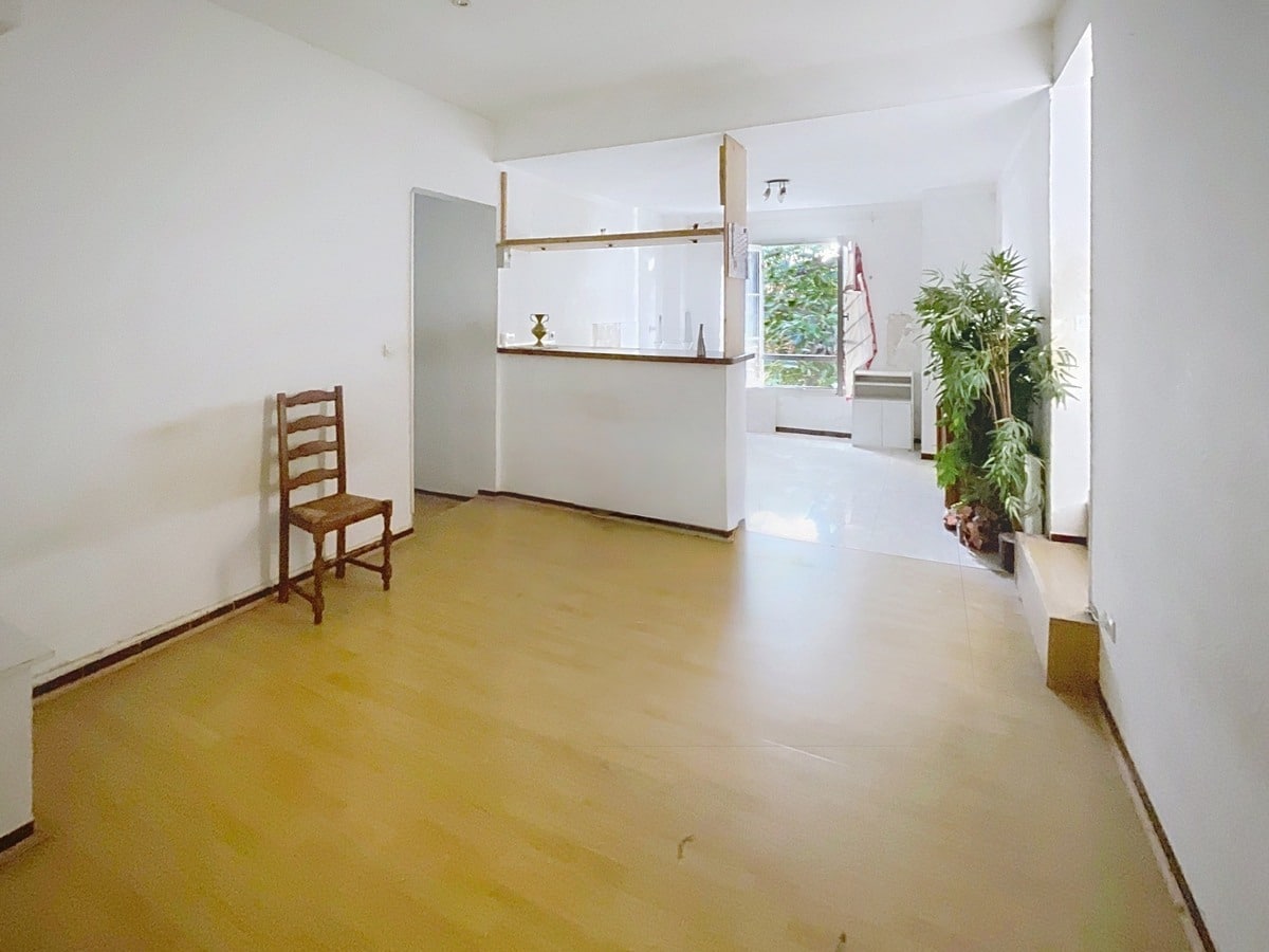 Séjour vide avec parquet clair et cuisine ouverte séparée par un bar en bois avec étagères. Un escalier rouge est visible à droite près d'une grande plante verte et d'une fenêtre donnant sur la végétation extérieure.