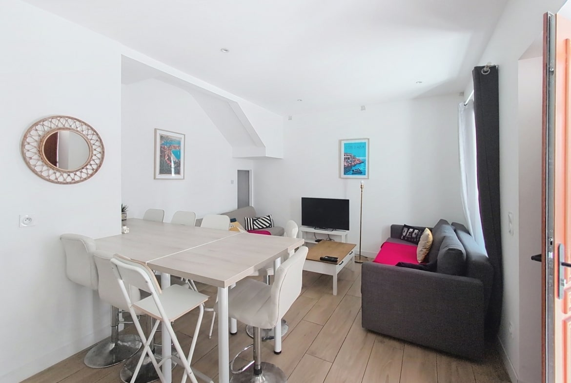 Pièce de vie ouverte avec table haute blanche entourée de chaises et tabourets assortis. Deux canapés gris avec plaids roses encadrent un meuble TV en bois clair. Des affiches décoratives représentant la Côte d'Azur ornent les murs blancs.