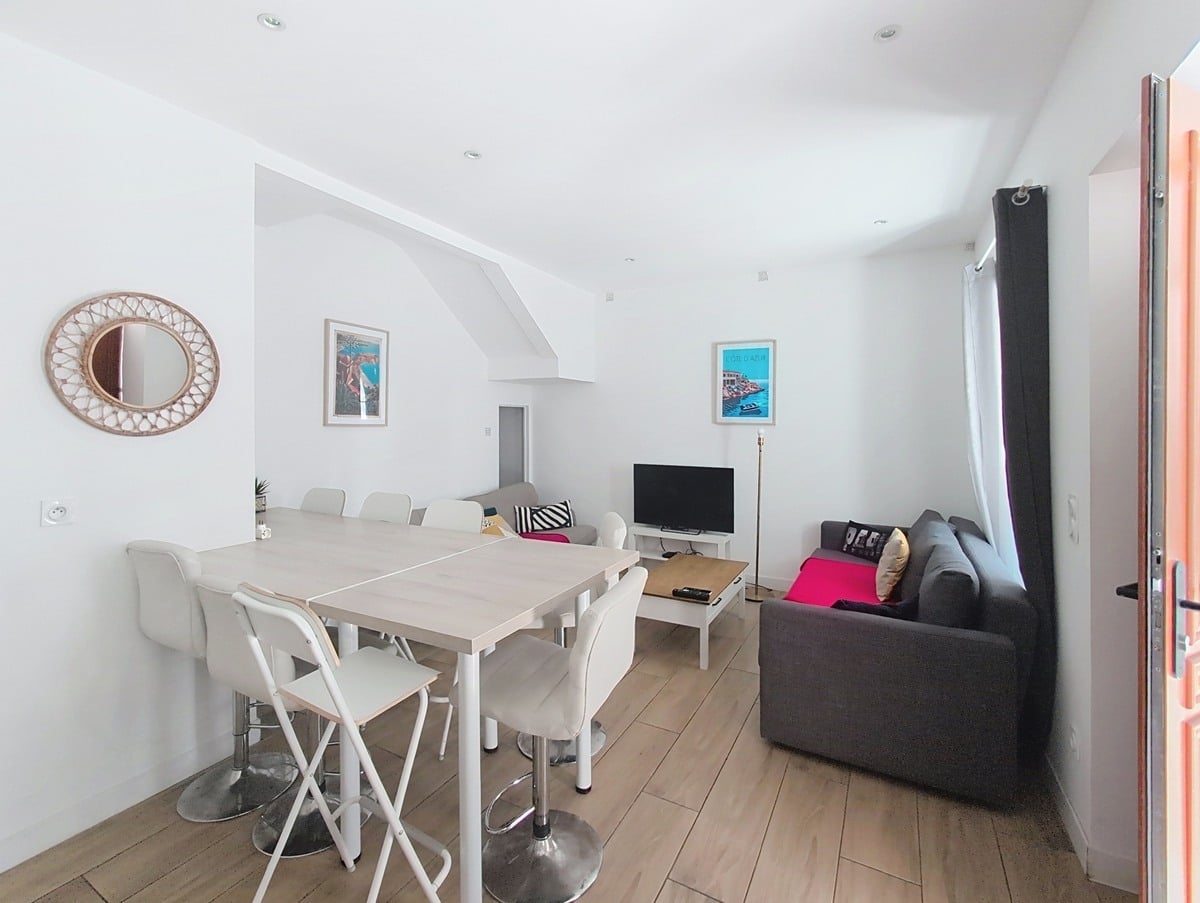 Pièce de vie ouverte avec table haute blanche entourée de chaises et tabourets assortis. Deux canapés gris avec plaids roses encadrent un meuble TV en bois clair. Des affiches décoratives représentant la Côte d'Azur ornent les murs blancs.