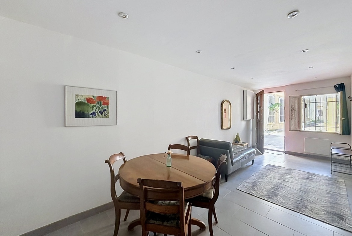 Séjour ouvert avec table ronde en bois et quatre chaises, canapé gris et tapis dans le prolongement. Murs blancs, carrelage gris au sol, tableau floral au mur et porte-fenêtre ouvrant sur la rue avec grilles en fer forgé.