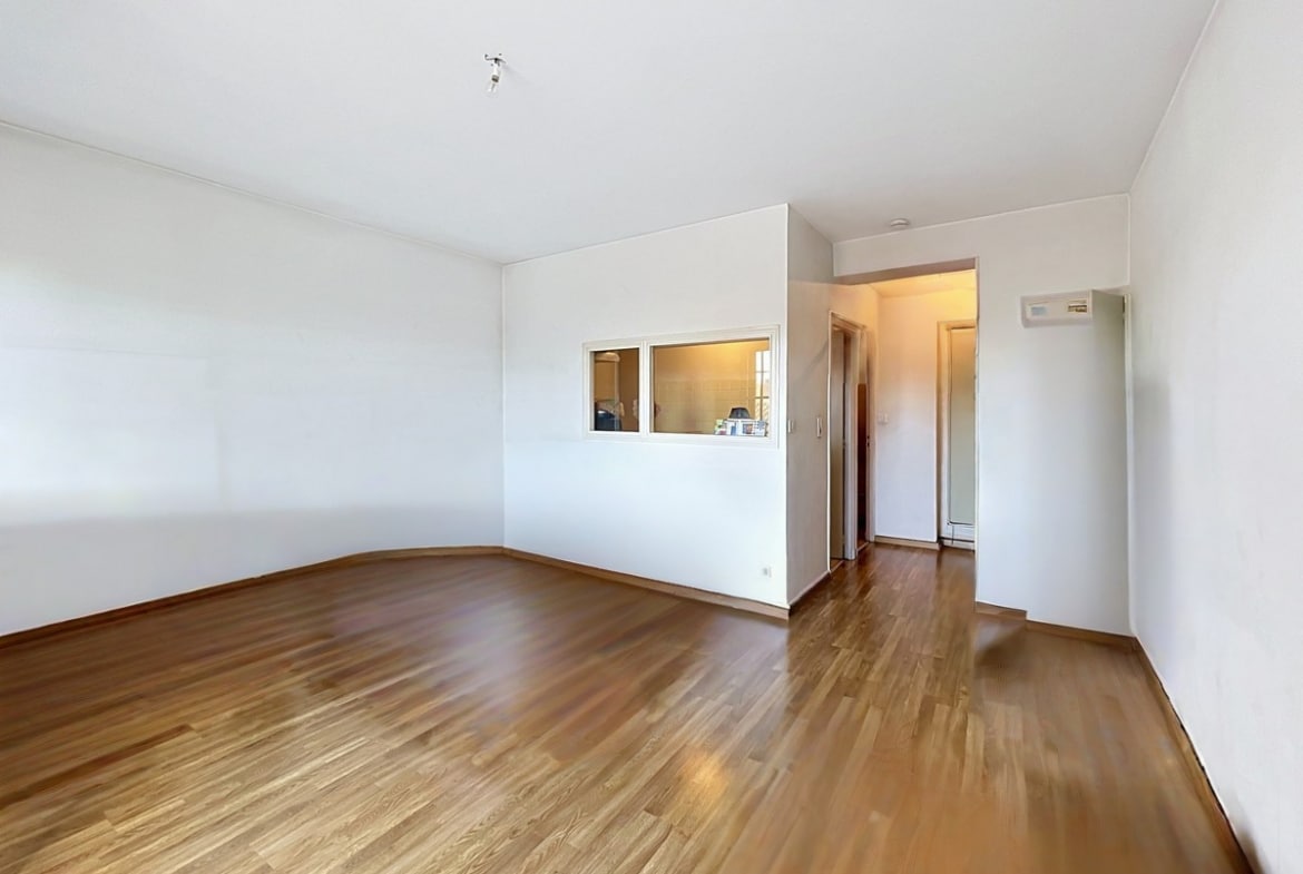Pièce de vie vide avec parquet foncé et murs blancs, passe-plat vitré donnant sur la cuisine. Couloir menant vers une entrée avec porte en bois, interphone visible sur le mur droit.