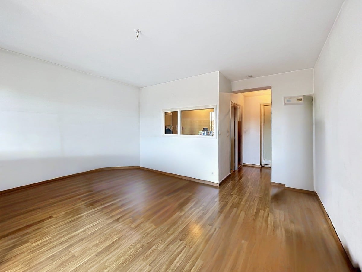Pièce de vie vide avec parquet foncé et murs blancs, passe-plat vitré donnant sur la cuisine. Couloir menant vers une entrée avec porte en bois, interphone visible sur le mur droit.