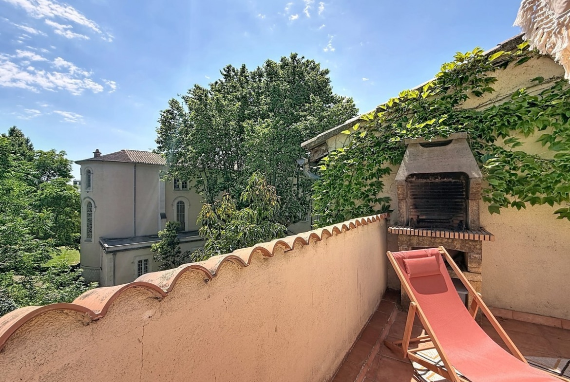 Terrasse carrelée en terre cuite avec muret surmonté de tuiles canal, barbecue en pierre intégré et transat rose. Vue dégagée sur grands arbres et bâtisse ancienne en arrière-plan, mur couvert de vigne vierge.
