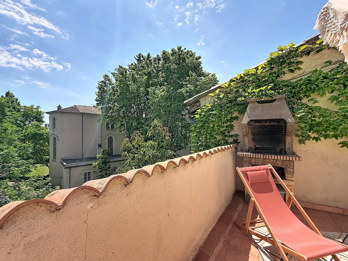Terrasse carrelée en terre cuite avec muret surmonté de tuiles canal, barbecue en pierre intégré et transat rose. Vue dégagée sur grands arbres et bâtisse ancienne en arrière-plan, mur couvert de vigne vierge.