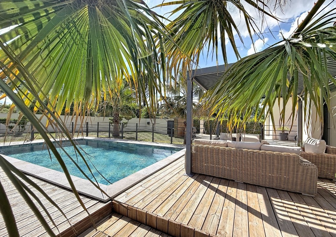 Terrasse en lames de bois naturel entourant une piscine aux eaux turquoise avec margelles en pierre claire. Canapé d'angle en résine tressée beige avec coussins blancs installé sous une pergola à lames orientables, encadré par des palmiers aux feuilles retombantes.