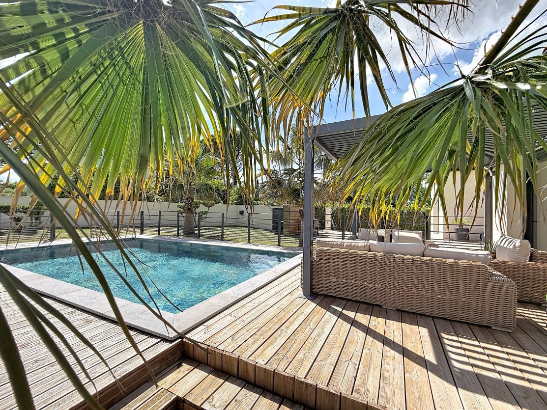 Terrasse en lames de bois naturel entourant une piscine aux eaux turquoise avec margelles en pierre claire. Canapé d'angle en résine tressée beige avec coussins blancs installé sous une pergola à lames orientables, encadré par des palmiers aux feuilles retombantes.