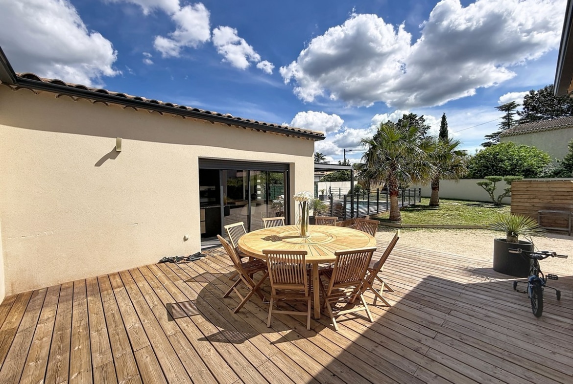 Terrasse en lames de bois naturel équipée d'une table ronde en teck avec huit chaises assorties. Vue sur le jardin avec palmier, pelouse et pergola bioclimatique sous un ciel partiellement nuageux.