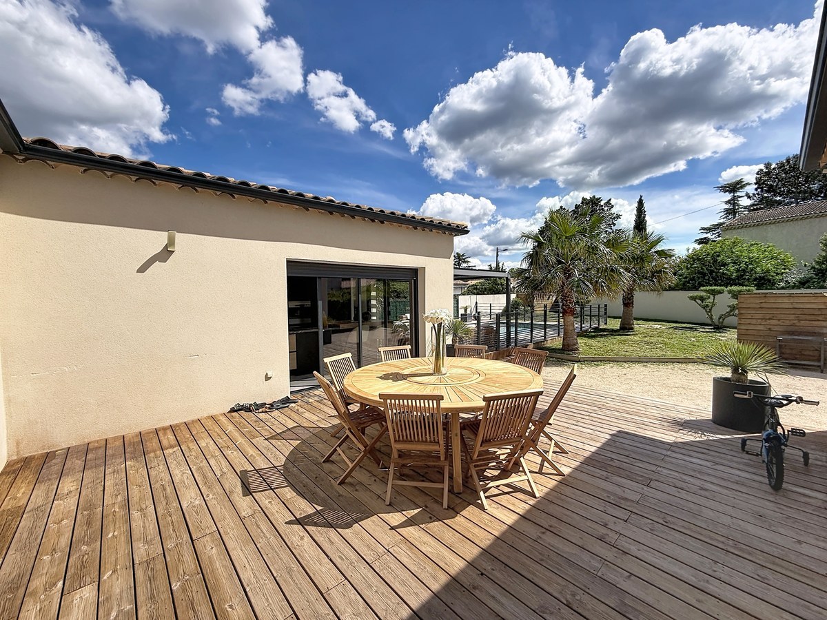 Terrasse en lames de bois naturel équipée d'une table ronde en teck avec huit chaises assorties. Vue sur le jardin avec palmier, pelouse et pergola bioclimatique sous un ciel partiellement nuageux.