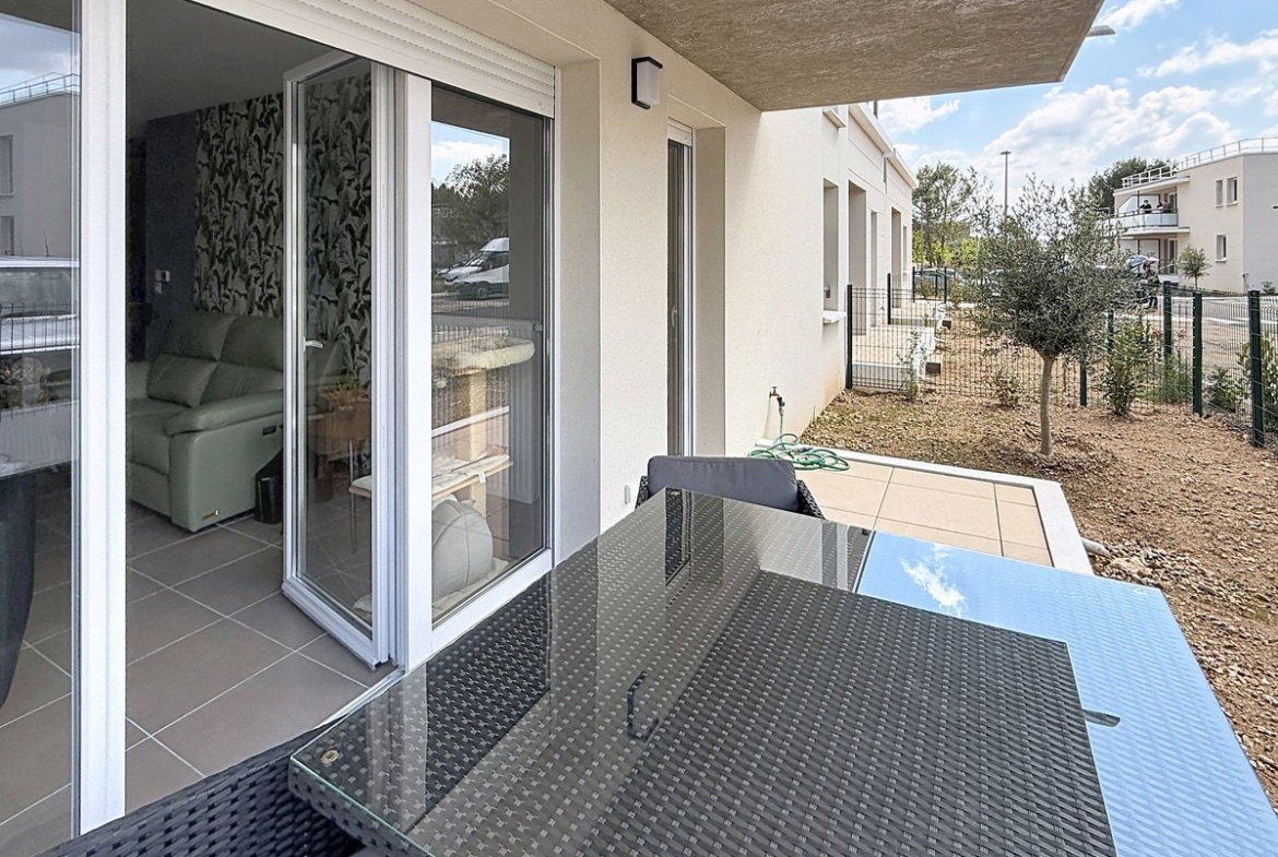 Terrasse couverte avec table et chaises en résine tressée noire, vue sur jardin privatif avec olivier planté. Baie vitrée coulissante donnant sur le salon avec papier peint végétal visible.