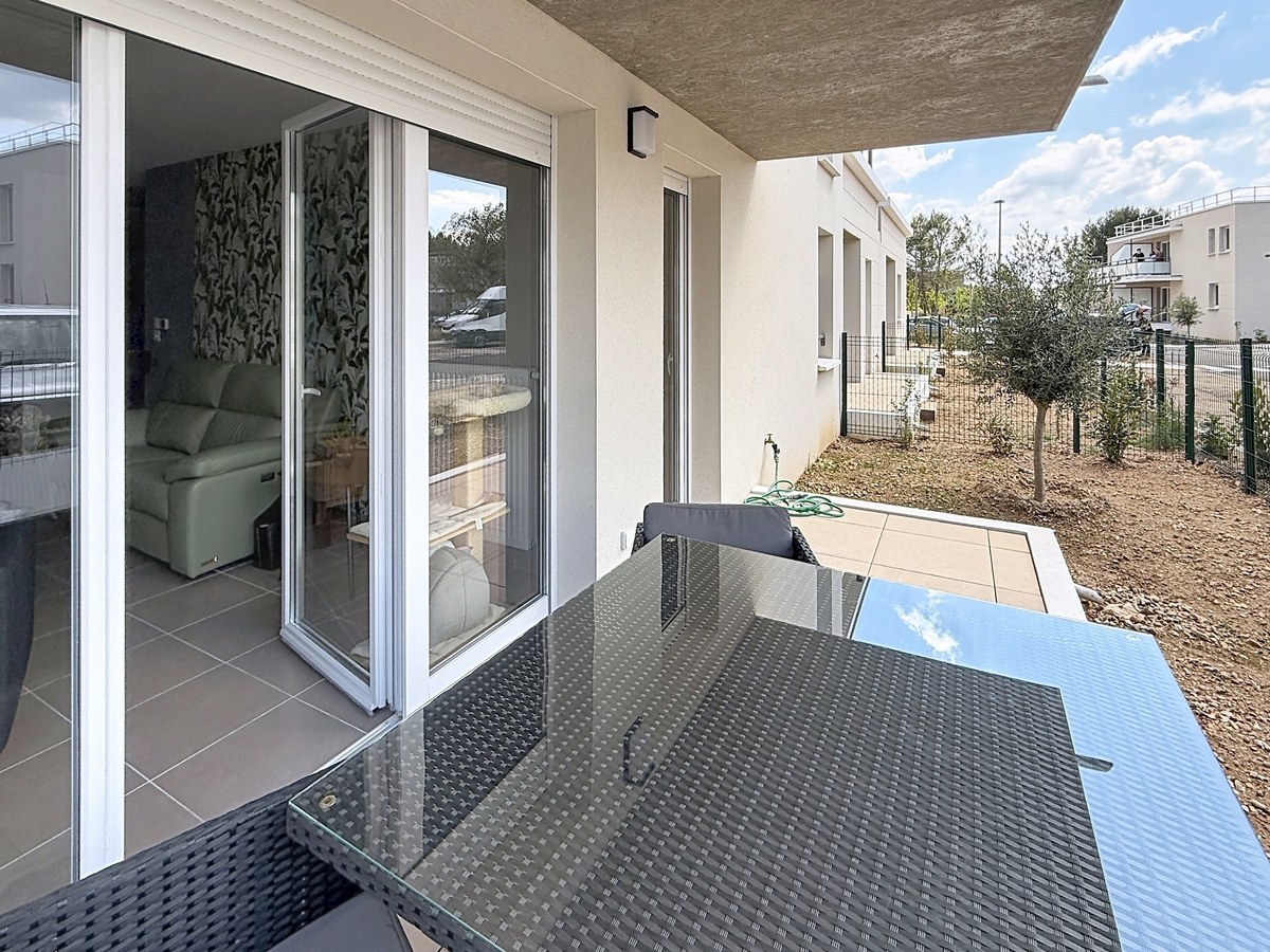 Terrasse couverte avec table et chaises en résine tressée noire, vue sur jardin privatif avec olivier planté. Baie vitrée coulissante donnant sur le salon avec papier peint végétal visible.