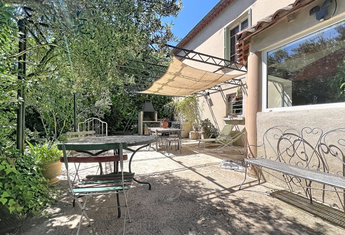 Terrasse dallée avec mobilier de jardin en fer forgé, pergola métallique équipée d'une voile d'ombrage beige et barbecue en pierre. Olivier et végétation méditerranéenne entourent l'espace, grande baie vitrée donnant sur l'intérieur.