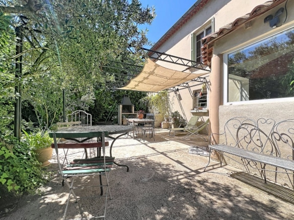 Terrasse dallée avec mobilier de jardin en fer forgé, pergola métallique équipée d'une voile d'ombrage beige et barbecue en pierre. Olivier et végétation méditerranéenne entourent l'espace, grande baie vitrée donnant sur l'intérieur.