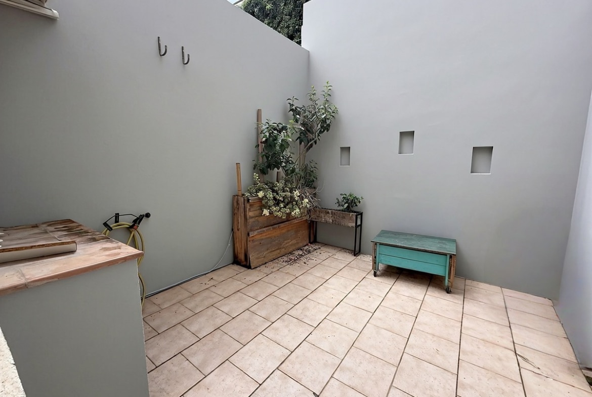 Terrasse intérieure avec sol carrelé beige entourée de murs gris. Des jardinières en bois rustique contiennent des plantes vertes, un banc turquoise sur roulettes est placé contre le mur percé de petites ouvertures carrées.