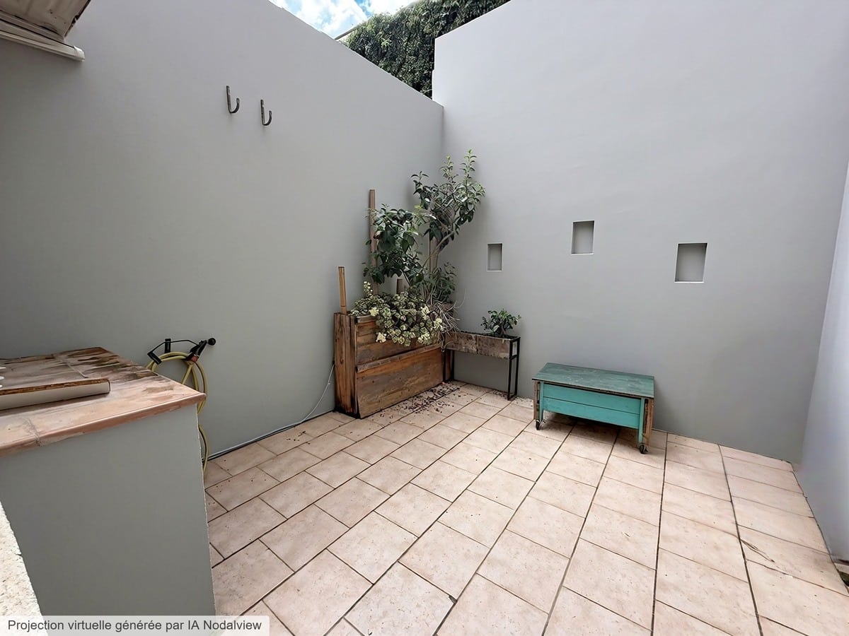 Terrasse intérieure avec sol carrelé beige entourée de murs gris. Des jardinières en bois rustique contiennent des plantes vertes, un banc turquoise sur roulettes est placé contre le mur percé de petites ouvertures carrées.