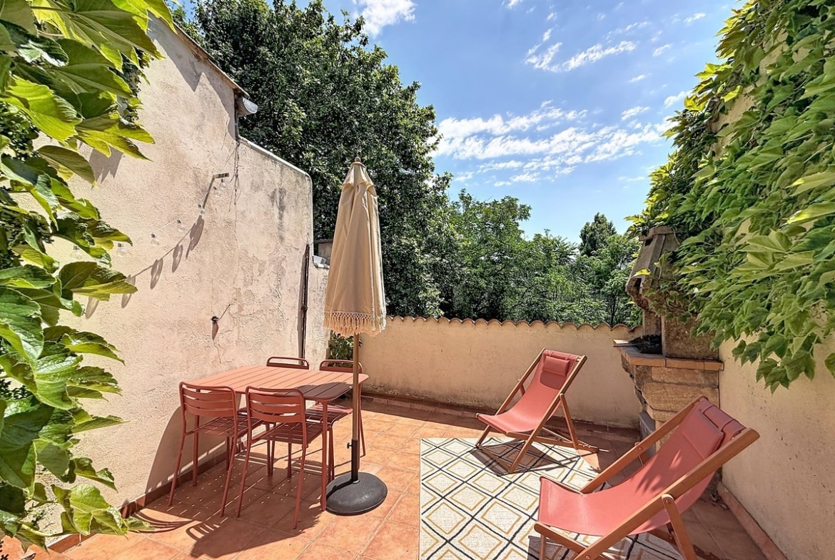 Terrasse avec table et chaises en métal couleur terracotta, parasol beige à franges et deux transats roses. Sol en carreaux de terre cuite, murs recouverts de vigne vierge, vue sur cimes d'arbres et ciel bleu.