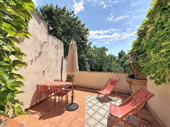 Terrasse avec table et chaises en métal couleur terracotta, parasol beige à franges et deux transats roses. Sol en carreaux de terre cuite, murs recouverts de vigne vierge, vue sur cimes d'arbres et ciel bleu.