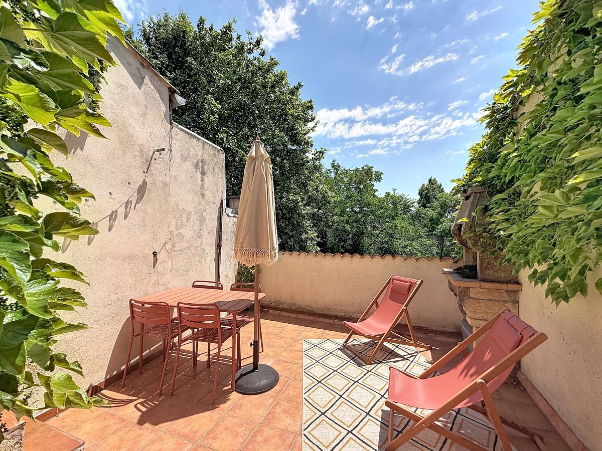 Terrasse avec table et chaises en métal couleur terracotta, parasol beige à franges et deux transats roses. Sol en carreaux de terre cuite, murs recouverts de vigne vierge, vue sur cimes d'arbres et ciel bleu.