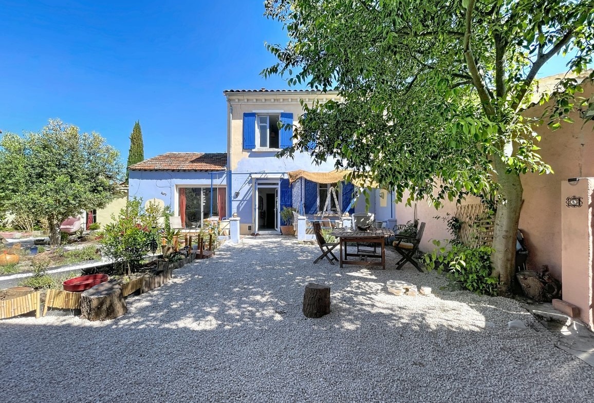 Façade de maison provençale à deux niveaux avec volets bleus et enduit blanc. Cour gravillonnée avec table en bois, olivier, cyprès et bassin décoratif. Voile d'ombrage beige tendu devant l'entrée sous un ciel bleu.