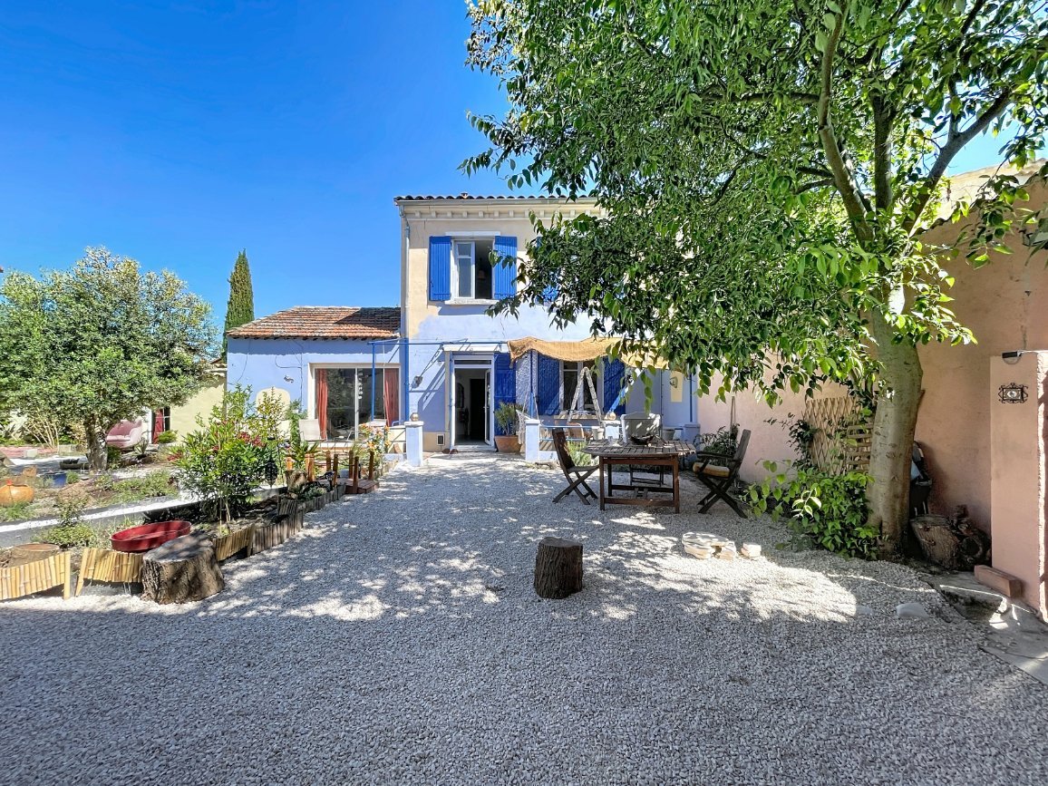 Façade de maison provençale à deux niveaux avec volets bleus et enduit blanc. Cour gravillonnée avec table en bois, olivier, cyprès et bassin décoratif. Voile d'ombrage beige tendu devant l'entrée sous un ciel bleu.