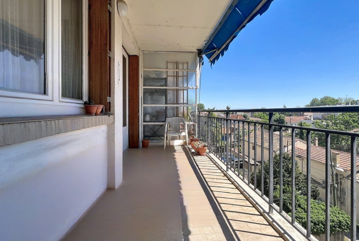 Long balcon étroit avec carrelage beige et garde-corps métallique noir donnant sur une vue dégagée des toits en tuiles. Store banne bleu déployé, chaises en plastique blanc et pots de fleurs en terre cuite disposés le long de la façade.