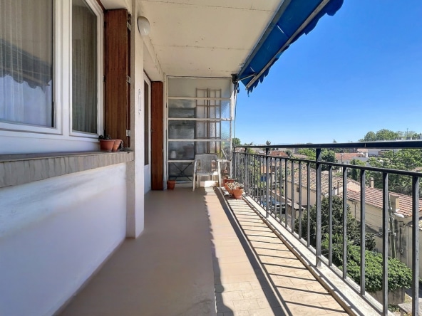 Long balcon étroit avec carrelage beige et garde-corps métallique noir donnant sur une vue dégagée des toits en tuiles. Store banne bleu déployé, chaises en plastique blanc et pots de fleurs en terre cuite disposés le long de la façade.