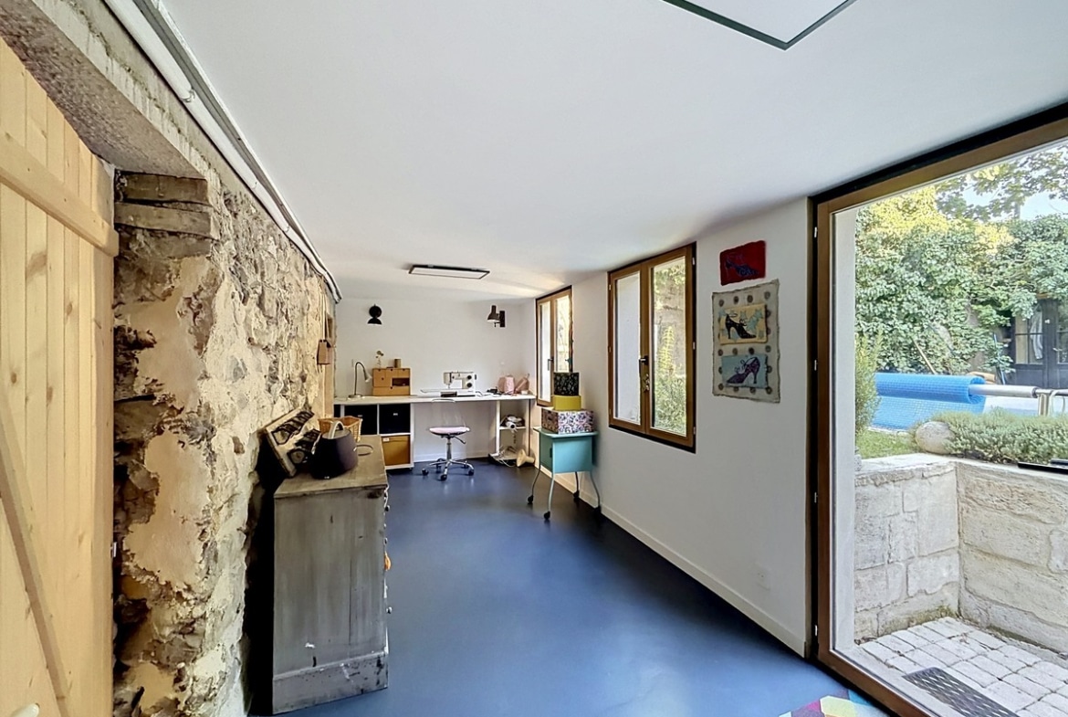 Espace atelier avec sol résine bleu, mur en pierre apparente et sauna en bois clair. Bureau blanc avec machine à coudre, grande porte-fenêtre ouvrant sur jardin et piscine, plafond blanc avec éclairage intégré.