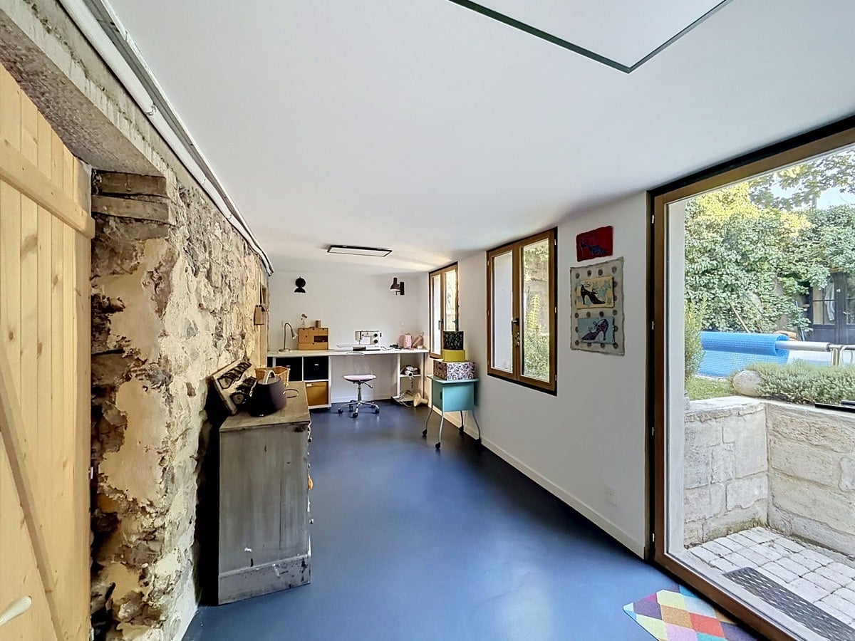 Espace atelier avec sol résine bleu, mur en pierre apparente et sauna en bois clair. Bureau blanc avec machine à coudre, grande porte-fenêtre ouvrant sur jardin et piscine, plafond blanc avec éclairage intégré.