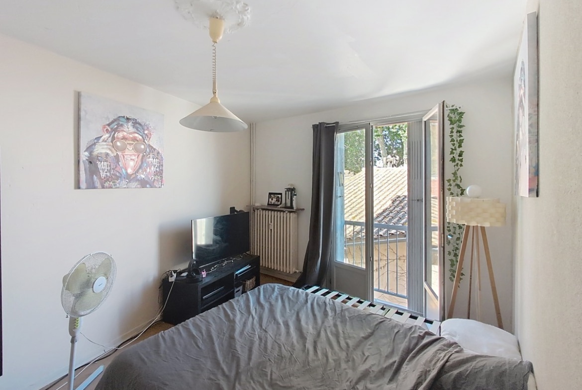 Chambre avec lit double recouvert d'un drap gris, meuble TV noir, ventilateur sur pied et lampadaire tripode en bois. Porte-fenêtre avec balcon donnant sur vue des toits, rosace décorative au plafond et tableaux colorés aux murs.